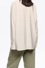 Camisa de corte recto en color beige claro, confeccionada en tejido fantasía con minibrillantes distribuidos por toda la prenda. Presenta cuello camisero y mangas largas con puño abotonado.