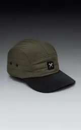 Gorra de estilo 5 paneles, color verde militar, con corona acolchada y visera negra. Presenta un pequeño parche negro con un logo blanco en forma de tijeras cruzadas.