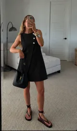 Vestido corto negro sin mangas, con cuello redondo y diseño cruzado decorado con botones claros en el lateral.