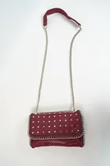 Cartera color bordó con solapa decorada con tachas en forma de estrellas plateadas. Tiene una correa de cadena plateada y un asa superior de tela bordó.