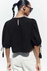 Blusa corta negra con mangas abullonadas y bordados florales en contraste de color blanco. Presenta cuello redondo con puntilla, mangas ajustables con cordones y borlas, y cierre en la espalda con abertura y botón oculto.