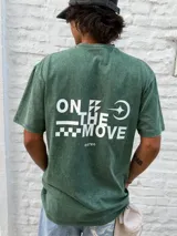 Remera de algodón color verde oscuro con efecto gastado (acid wash). Presenta un estampado minimalista en el pecho con el texto "ON THE MOVE" y un patrón de cuadros blancos a la derecha.