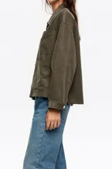Chaqueta de corte recto y holgado en color verde oliva, confeccionada en tejido de algodón con textura. Presenta cuello camisero, cierre frontal con botones metálicos y dos bolsillos frontales de parche.
