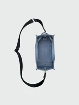 Bolso tipo tote de lona color beige de la marca Marc Jacobs. Presenta el texto 'THE TOTE BAG' estampado en el frente en color blanco, dos asas de mano superiores y una correa de hombro ajustable y removible en color negro.