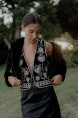 Chaleco negro con escote en V profundo, confeccionado en tela con bordados florales en hilo plateado. Presenta un diseño estructurado con ribetes plateados en los bordes y cierre frontal.