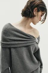 Jersey de punto color gris oscuro, con escote recto tipo off-shoulder, manga larga y detalle de aplique metálico en el hombro. Presenta acabados en rib en puños y ruedo.