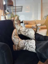 Botas de caña media con estampado de piel de serpiente en tonos beige y marrón. Diseño estilo cowboy con punta cuadrada, detalle de hebilla y apliques metálicos.