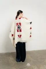 Ruana tejida en color crudo con diseño geométrico inspirado en motivos Navajo en tonos bordó y negro. Presenta terminaciones con flecos en el ruedo y un pequeño aplique de cuero en la parte inferior.