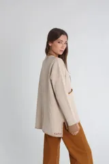 Saco de punto color beige con textura acanalada, cuello en V y cierre frontal con botones. Presenta bolsillos delanteros con bordes en color marrón y un pequeño detalle bordado en rojo sobre uno de los bolsillos.