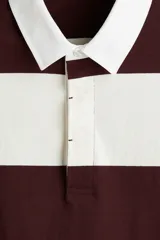 Remera tipo polo de manga larga con diseño a rayas horizontales en color bordó y blanco. Presenta cuello inglés, tapeta con botones ocultos y puños acanalados.
