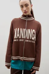 Campera tejida de punto color marrón con cierre frontal completo, cuello alto y detalles de rayas blancas en cuello, puños y ruedo. Presenta el logo de la marca estampado en el frente.