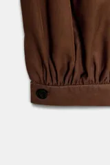 Pantalón de tiro alto en color beige, con diseño de pierna holgada y puños elásticos con cierre de botón en el bajo. Presenta cierre frontal con cremallera y doble botón en la cintura, además de bolsillos laterales.
