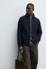 Chaqueta de punto con textura acanalada, cuello subido y cierre frontal completo con cremallera metálica. Presenta un corte relajado y acabados en rib en puños y ruedo.