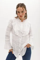 Camisa blanca de algodón con diseño romántico, cuello alto con volados, cierre frontal con botones nacarados y mangas largas con puños acampanados. Presenta detalles de encaje y franjas horizontales en toda la prenda.