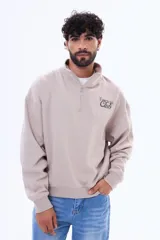 Buzo de felpa color beige con cuello alto, cierre frontal parcial y logo bordado en el pecho.