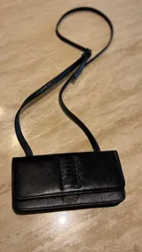 Cartera tipo bandolera de cuero negro, de diseño rectangular y compacto. Presenta una solapa frontal con detalle de costuras cruzadas decorativas y un pequeño aplique metálico con logo. Incluye correa larga regulable y cierre mediante botón imantado.