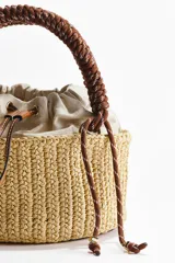 Cartera tipo tote en paja sintética trenzada, con estructura cilíndrica. Cuenta con un bolso interior de lona color beige con cordón ajustable, asa superior trenzada de cuerda marrón y detalles en ecuero.