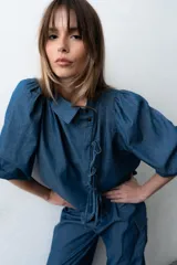 Blusa de denim liviano de corte asimétrico en tono azul, con mangas abullonadas, cuello redondo y cierre lateral con lazos para atar y botones.