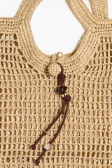 Cartera tote de paja trenzada con diseño calado, asas de hombro y cierre con cordón y cuentas decorativas.