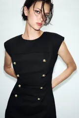Vestido corto de estilo militar en color negro, con cuello redondo, hombreras y manga sisa. Presenta un diseño frontal con falsos botones dorados dispuestos en filas horizontales. Cierre lateral con cremallera oculta y abertura en la espalda con botón.