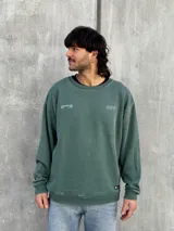 Buzo verde de algodón con cuello redondo, mangas largas y puños acanalados. Presenta un pequeño estampado en el frente y un logo geométrico en el pecho. Corte holgado.