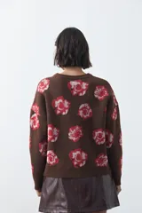 Cardigan de tejido jacquard color marrón con estampado floral en tonos rojo y rosado. Presenta cuello redondo, cierre frontal con botones y terminaciones acanaladas en puños y ruedo.