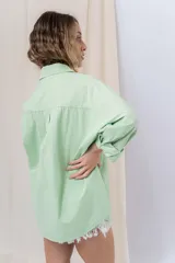 Camisa verde pastel de algodón, corte oversize, con cuello camisero, cierre con botones, mangas largas con puños con volados de encaje blanco, dos bolsillos frontales con solapa y ruedo irregular. 