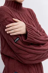 Sweater de punto con diseño de trenzas, cuello alto y mangas largas con puños acampanados. Presenta un corte holgado con terminación asimétrica en el ruedo.