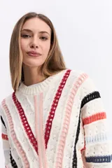 Buzo de tejido calado con diseño de rayas horizontales y verticales en colores blanco, rojo, negro y rosa. Presenta cuello redondo, mangas largas y un corte corto tipo crop.