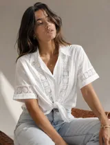 Camisa blanca de manga corta con cuello camisero, detalles de encaje aplicado en el frente y en las mangas, y nudo en la parte inferior delantera.