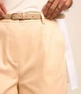 Short de vestir color beige, con cintura alta, pliegues frontales, cinturón de cuero sintético con hebilla metálica y cierre frontal con botón y cremallera.