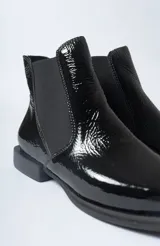 Botín Chelsea de mujer, color negro charol, con elástico lateral y punta cuadrada. Posee un taco bajo y plano.