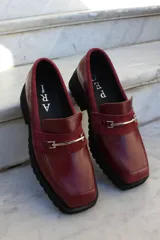 Mocasín de cuero vacuno color bordó con suela track negra de goma. Presenta un diseño de punta cuadrada con un herraje metálico plateado sobre una banda de gamuza en la capellada.