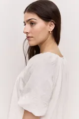Blusa blanca de manga corta, con escote redondo y cierre frontal con tres botones. Presenta pliegues frontales y diseño liviano.