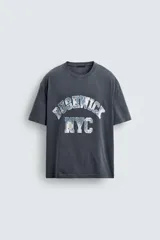 Camiseta de corte boxy fit con cuello redondo y manga corta. Presenta un diseño con efecto lavado y estampado gráfico frontal con tipografía universitaria.