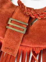 Cartera de cuero gamuza color naranja con diseño de media luna, decorada con flecos largos en los laterales. Presenta un cierre metálico superior, un herraje metálico distintivo en forma de S en el frente y un asa corta trenzada.
