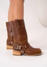 Bota de caña media confeccionada en cuero vacuno color suela, con diseño estilo western. Presenta una tira con hebilla metálica y estribo desmontable en el tobillo, puntera redonda y taco cuadrado de madera con suela de caucho.