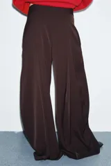 Pantalón de tiro medio con pierna ancha, confeccionado en tela de color marrón oscuro. Presenta pliegues frontales decorativos, bolsillos laterales ocultos en la costura y cierre frontal con cremallera, botón y ganchos metálicos.