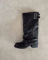 Bota alta de caña arrugada, color negro, confeccionada en cuero vegano. Presenta un taco bajo y hebillas decorativas en la parte superior de la caña.