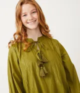 Vestido corto de manga larga en color verde oliva, confeccionado en tejido plano con hilos metalizados a contraste. Presenta cuello redondo con lazo decorativo y flecos, mangas largas con puños elásticos y detalle de volados en los puños.