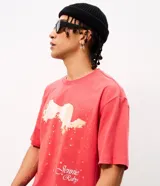Remera de corte boxy en color rojo, con cuello redondo y mangas cortas. Presenta una estampa frontal con una ilustración fotográfica y la firma Jennie Ruby en tipografía cursiva.