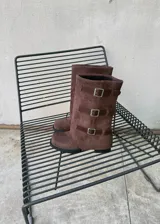 Botas de caña alta de gamuza marrón con puntera cuadrada, suela plana y tres tiras laterales con hebillas metálicas.