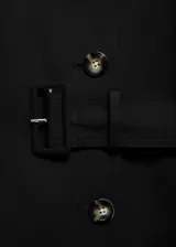 Trench coat negro de diseño recto y largo, con cuello de solapa, trabillas en los hombros y en las mangas, y cierre frontal con botones. Incluye bolsillos laterales y abertura posterior en el bajo.