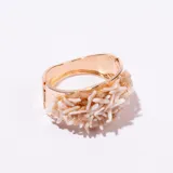 Brazalete tipo esclava de metal dorado, con un diseño ancho y textura acanalada en la parte interna. La parte externa está decorada con múltiples piezas irregulares de color blanco hueso o coral claro, sujetas con alambre fino dorado, simulando ramas o corales.