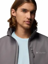 Chaqueta Columbia modelo Ascender Soft Shell, color gris, con cierre frontal completo, cuello alto con protector de barbilla, bolsillo en el pecho con cierre y bolsillos laterales. Cuenta con puños ajustables y tecnología resistente al agua y al viento.