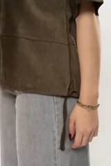 Remera de gamuza color rojo oscuro, con cuello redondo, mangas cortas y detalle de cortes verticales y horizontales que definen la silueta. Presenta cordones ajustables en el ruedo inferior.