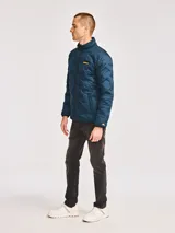 Chaqueta acolchada de peso medio en color azul petróleo, con cierre frontal de cremallera, cuello alto y parche de silicona con el logotipo de la marca en el pecho. Cuenta con bolsillos laterales y ribetes elásticos en puños y dobladillo.