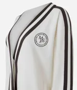 Cardigan deportivo de corte oversized, color blanco con detalles en marrón oscuro. Presenta escote en V, cierre frontal con botones a presión, mangas largas con franjas laterales y puños acanalados. Incluye un parche circular con letras en el pecho.