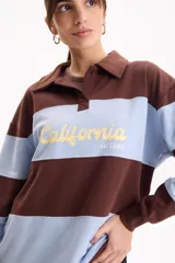 Buzo deportivo sin friza a rayas anchas en tonos blanco y tostado, con cuello polo, escote en V y estampa frontal con la inscripción "California est.1969" en color fucsia. Presenta un corte semi oversize y mangas largas.