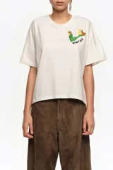 Remera de manga corta color crudo, con cuello redondo y diseño holgado. Presenta un bordado en el pecho con motivo verde y amarillo y la inscripción de la marca.